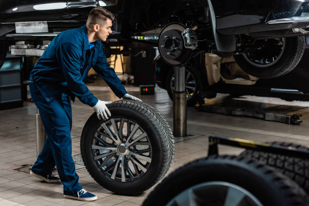 giovane meccanico che tiene ruota dell'auto vicino auto rialzata in officina - work glove immagine foto e immagini stock