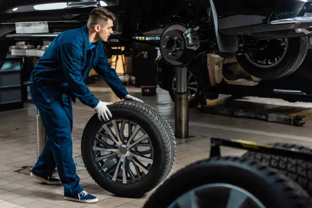 Photo of young mechanic holding car wheel near raised car in workshop