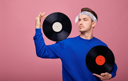 Retro style calm hippie guy in bandana in dark blue sweater stands on pink background with one black vinyl on the shoulder, and with the second record in the other hand at chest level. Looks sideway.