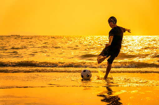 Kid Playing Soccer Beach Summer Concept . A boy playing football by the sea during sunset. Tourists or exercise people are breathing good air.