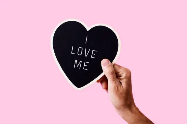 closeup of a man holding a black heart-shaped sign with the text I love me written in it, on a pink background