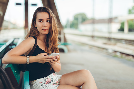 Young woman waiting for the train and using phone on a railway station.