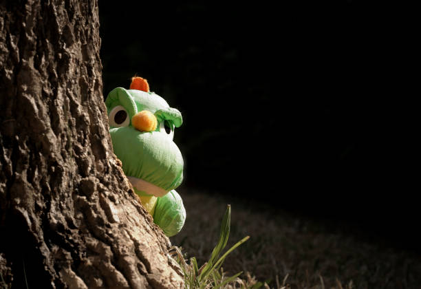 Sunlight on little green dinosaur plush doll in peek gesture behind a big tree trunk in the garden, close up with copy space