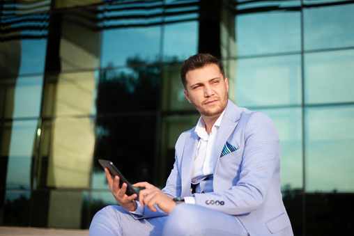 Low angle portrait of young businessman using digital tablet looking away with blank facial expression against office exterior. Horizontal shot.