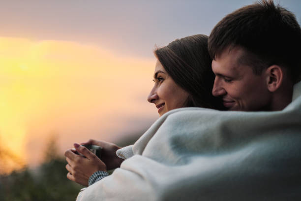 A couple in love sits on the riverbank at sunset. They cuddle, roll into a plaid and drink tea from a metal hiking mug. stock photo