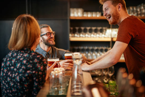 cheerful friends leaning on bar counter, drinking beer and chatting with bartender. night out. - irish culture beer drinking pub imagens e fotografias de stock
