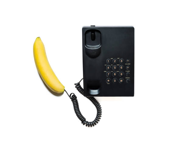 Creative photo of a simple modern black push-button telephone with a bright yellow banana instead of a handset close-up, isolated on a white background. stock photo