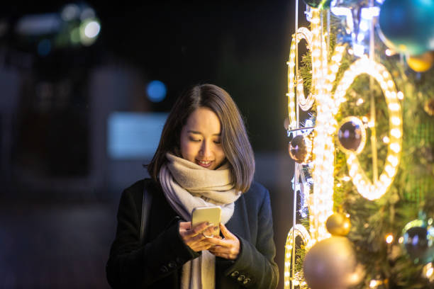 Young woman standing by Christmas lights and typing message stock photo
