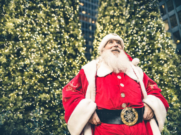 Santa Clause posing in front of large Christmas tree in big North American city. Long white beard, Red suit, boots and all. Ready for Christmas.
