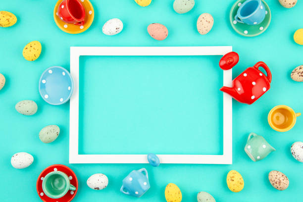 Toy tea cups and Easter eggs with white frame over turquoise paper background. Easter celebration banner, party invitation, greeting card concept. Flat lay, top view