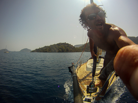 An action cam shot of an alone sailor while standing at the bow of a moving sailing boat in the mediterranean summer.