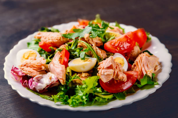 istock Fresh salad with fish, arugula, eggs,red pepper, lettuce, fresh sald leaves and tomato on a white plate on wooden table background 1195872583