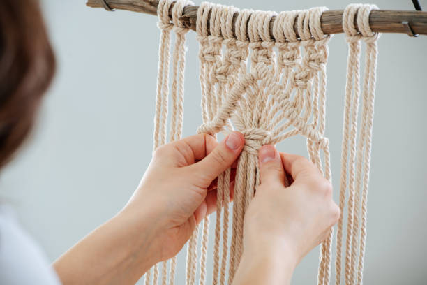 Craftswoman weving ropes, creating a macrame banner. from behind. Brunette woman working on a half-finished macrame piece, weaving ropes, making knots. Close up. Cropped. From behind. macrame photos stock pictures, royalty-free photos & images