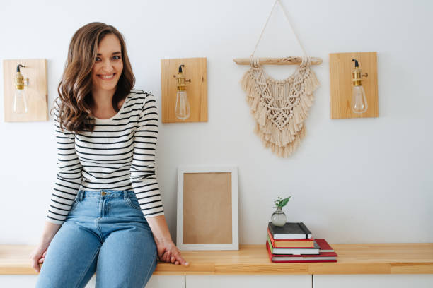 Young cheerful woman sitting on a shelf in a corridor, next wall lamps Young cheerful brunette woman with long and beautiful hair is sitting on a shelf in a corridor, next to wall lamps, stack of books and macrame tapestry. macrame photos stock pictures, royalty-free photos & images