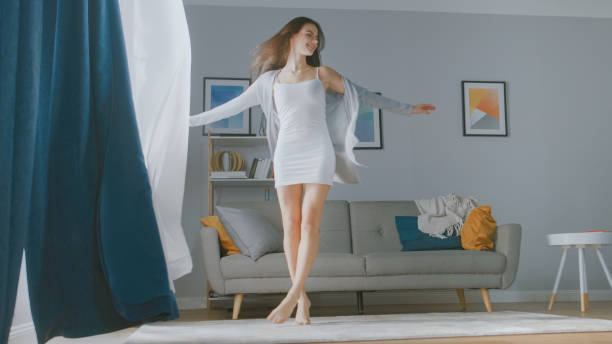 beautiful young brunette dances in the morning light illuminates her cozy living room. sensual girl rising early and greeting new day. - sunny apartment window sky imagens e fotografias de stock