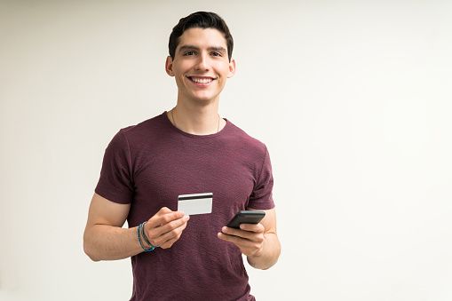 Happy attractive young male holding credit card and mobile phone against isolated background