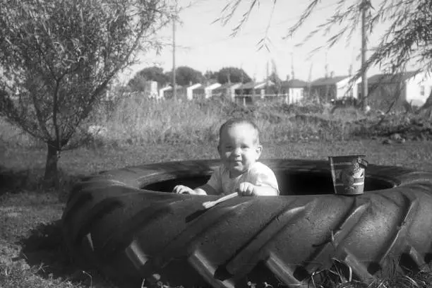 Baby playing in tractor tire sandbox in front of a row of new tract homes. Symbolic of post WW2 affluence and baby boom. Waterloo, Iowa, USA 1952. Scanned film.