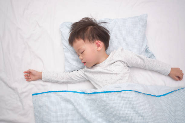 Cute little Asian 3 - 4 years old toddler boy kid in pajama taking a nap, sleeping on his back  on white bed sheet in bed stock photo