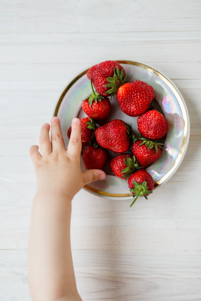 ręka dziecka z truskawką na rustykalnym tle, talerz truskawek. - juicy childhood colors red zdjęcia i obrazy z banku zdjęć