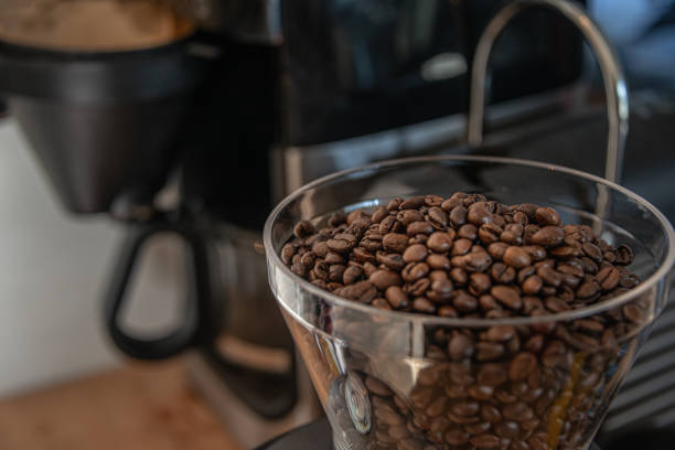 Transparent coffee grinder with coffee beans and filter coffee machine in background