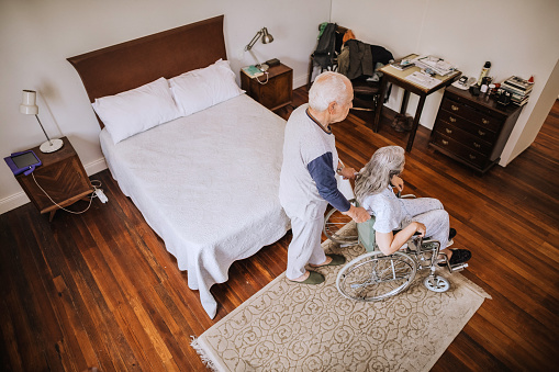 Every morning routine for senior couple. They are in pajamas in their bedroom. Husband helping his senior wife with wheelchair