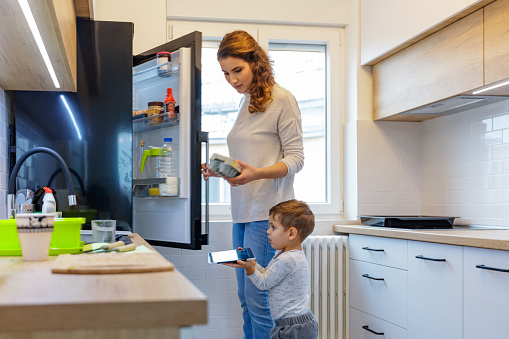 Millennial mother taking egg carton out of refrigerator, her cute 2,5 year old son is holding digital tablet
