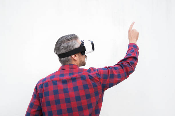 Back view of man in VR headset pointing with finger. Rear view of bearded man in checkered shirt using virtual reality headset on grey background. Technology concept