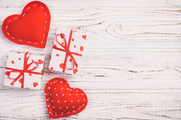 Valentine or other holiday handmade present in paper with red hearts and gifts box in holiday wrapper. Present box of gift on white wooden table top view with copy space, empty space for design. san valentín u otro regalo de vacaciones hecho a mano en papel con corazones rojos y caja de regalos en envoltorio de vacaciones. caja de regalo presente en la vista superior de la mesa de madera blanca con espacio de copia, espacio vacío para el diseño