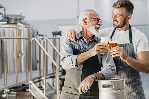Portrait of a senior man and his son in a craft brewery while cheering with beer