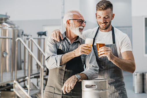 Portrait of a senior man and his son in a craft brewery while cheering with beer