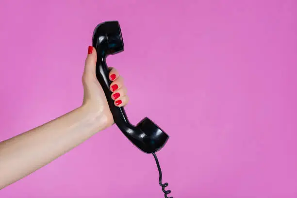 Beautiful young girl and gently hand holding old and retro telephone headset isolated on pink background in studio. Girl hands with red manicure on fingers. Minimalism and retro concept