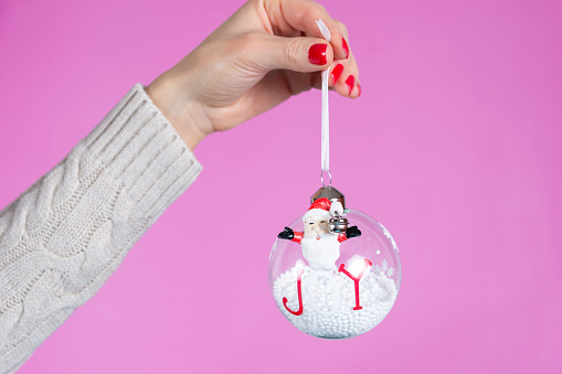 Beautiful young girl and gently hand holding ornament ball with snow isolated on pink background in studio. Female hands with red nails polish on fingers. Christmas or Valentines day holiday and minimalism concept