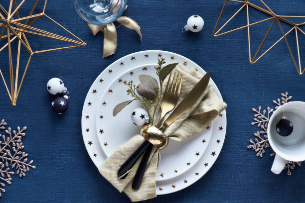 istock Christmas table setup with white plate and golden utensils and gilded decorations. White and black glass baubles with stars. Flat lay, top view on dark blue linen textile background. 1196100650
