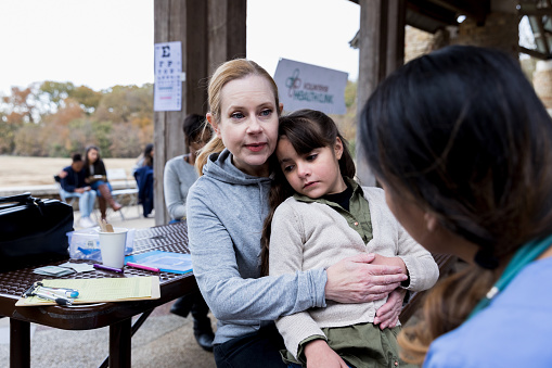 Concerned mid adult mom comforts her ill elementary age daughter while visiting an outdoor free clinic. The mom is talking with a female doctor or nurse.