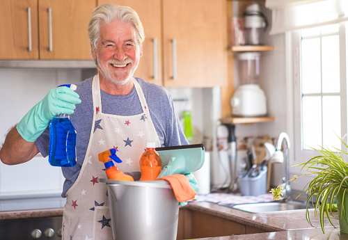 A senior smiling man ready to start housework. He holds a plastic bucket with cleaning items inside and a spray bottle in his hand. Only one people with green gloves