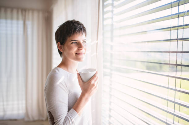 Portrait of young woman with coffee standing by window indoors at home. Portrait of young happy woman with coffee standing by window indoors at home. window blinds photos stock pictures, royalty-free photos & images