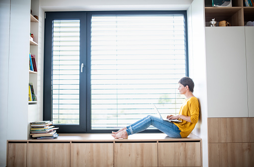 Side view of young woman with laptop sitting on window sill in bedroom indoors at home.