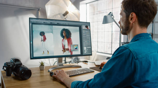 il fotografo professionista seduto alla sua scrivania utilizza il computer desktop in un photo studio retouches. dopo il servizio fotografico tocca le fotografie di una bella modella nera in un software di editing di immagini - editor foto e immagini stock