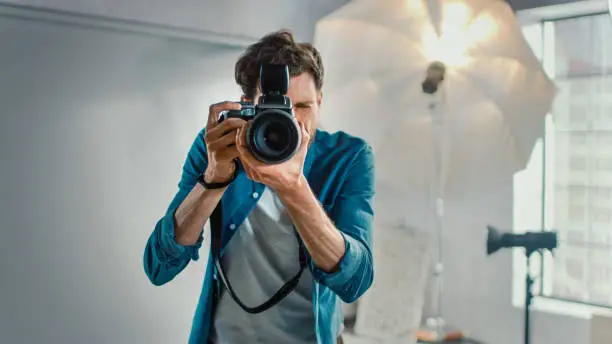In the Photo Studio with Professional Equipment: Portrait of the Famous Photographer Holding State of the Art Camera Taking Pictures with Softboxes Flashing in Background.