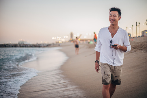 Hipster man walking on the beach and listening to music on the mobile phone