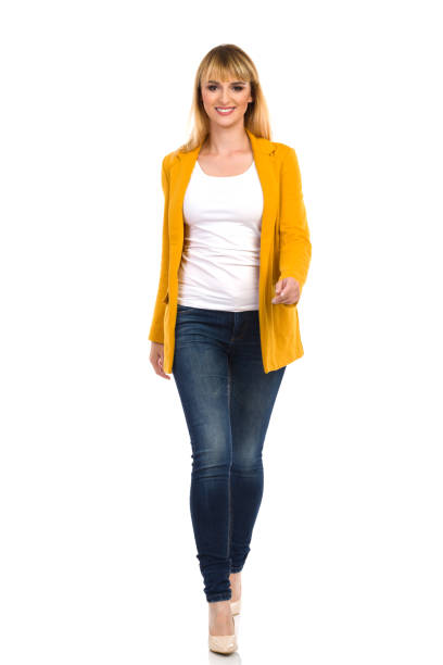 Confident Young Woman In Yellow Unbuttoned Jacket Is Walking Towards Camera. Front View. Confident smiling young woman in yellow unbuttoned jacket, blue jeans and high heels is walking towards camera. Front view. Full length studio shot isolated on white. approaching stock pictures, royalty-free photos & images