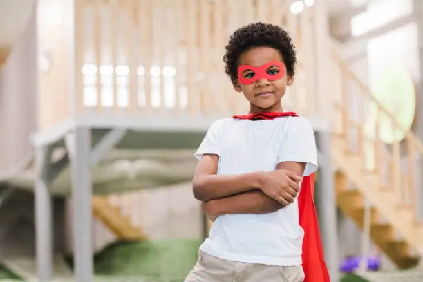 Cute African little boy in white casualwear and red mantle of superman looking at you while playing in kindergarten