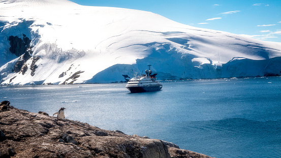 A scenic view of the Antarctic peninsula with a penguins in the. foreground and a small expedition ship in the background.  Approximately 40 thousand people will have visited the Antarctic peninsula in 2019.