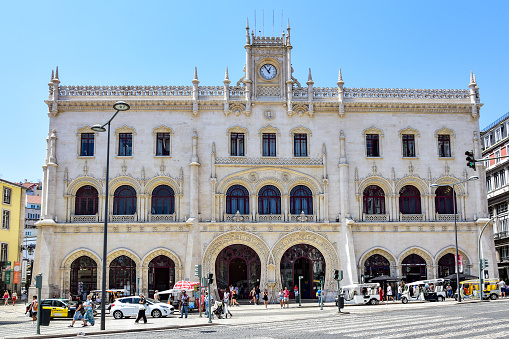 Lisbon, Portugal - Rocio Station (Rossio Train Station), a beautiful white building with horseshoe-shaped doors and a clock at the top, a street with cars and people in the summer afternoon.
