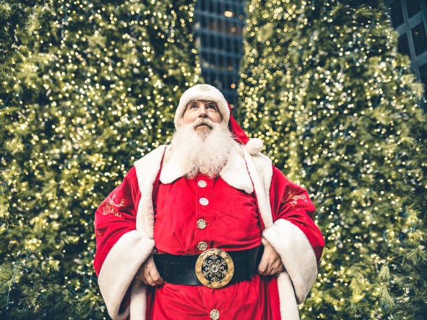 Santa Clause posing in front of large Christmas tree in big North American city. Long white beard, Red suit, boots and all. Ready for Christmas.