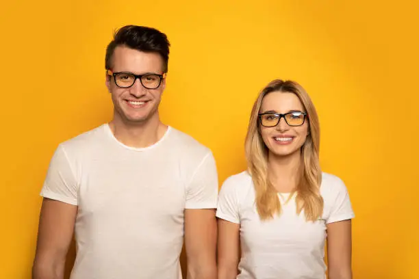 Close-up photo of a man and woman, posing in front of white t-shirts and glasses and smiling while looking in the camera.