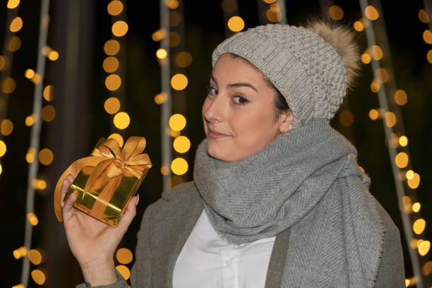 Close-up of a smiling woman holding a golden wrapped gift box while looking at the camera on an out of focus background. Shopping for gifts concept.