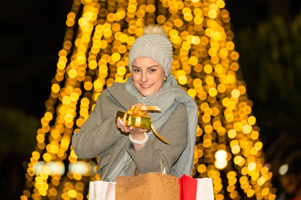 Smiling woman shows a golden wrapped gift box at the camera while holding several paper gift bags on an out of focus sparkly background. Shopping for gifts concept.