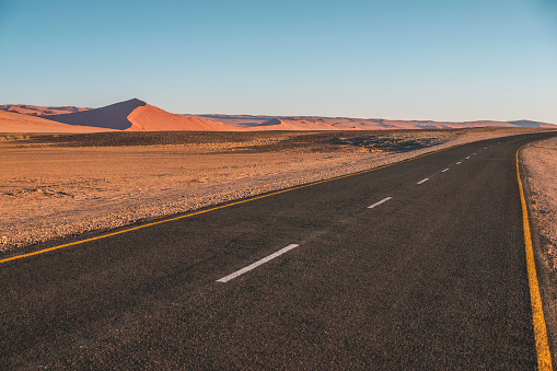 Empty Open Desert Road in arid vast Desert Landscape of the Sossusvlei Namib Desert of Namibia. Giant Sand Dunes roadside the Highway in the background. Roadside panorama under blue summer skyscape. Namibian Desert, Sossusvlei, Namibia, Southern Africa.