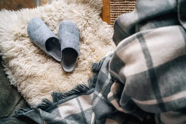 The pair of gray home slippers near the  bed on the white sheepskin in the cozy bedroom. Home sweet home concept top view image.